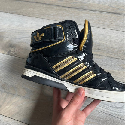 Adidas Space Diver Zip Pack Sneakers were released on November 18, 2012. The shoes feature a black upper with a glossy leather-like finish, gold detailing, and a white outsole and insole. The tongue has a large gold Trefoil logo with a gold outline, while the heel strap is printed with gold Adidas text. The sides feature three gold stripes designed as decorative zipper elements. A small white Adidas logo is also present on the upper.