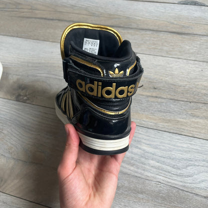Adidas Space Diver Zip Pack Sneakers were released on November 18, 2012. The shoes feature a black upper with a glossy leather-like finish, gold detailing, and a white outsole and insole. The tongue has a large gold Trefoil logo with a gold outline, while the heel strap is printed with gold Adidas text. The sides feature three gold stripes designed as decorative zipper elements. A small white Adidas logo is also present on the upper.