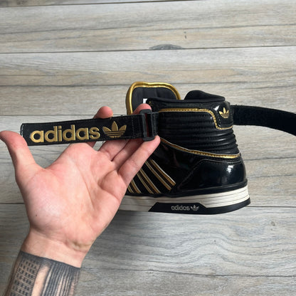 Adidas Space Diver Zip Pack Sneakers were released on November 18, 2012. The shoes feature a black upper with a glossy leather-like finish, gold detailing, and a white outsole and insole. The tongue has a large gold Trefoil logo with a gold outline, while the heel strap is printed with gold Adidas text. The sides feature three gold stripes designed as decorative zipper elements. A small white Adidas logo is also present on the upper.