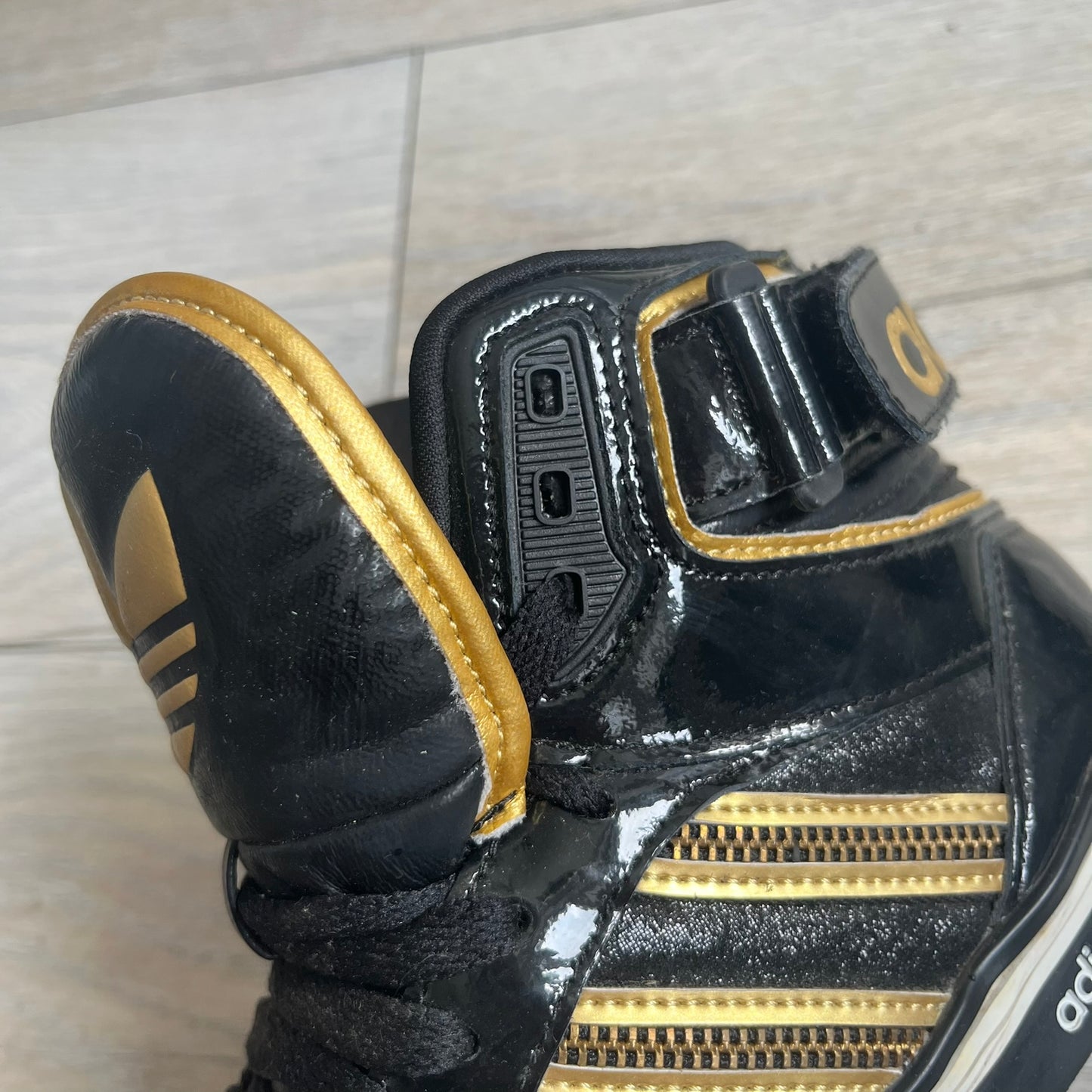 Adidas Space Diver Zip Pack Sneakers were released on November 18, 2012. The shoes feature a black upper with a glossy leather-like finish, gold detailing, and a white outsole and insole. The tongue has a large gold Trefoil logo with a gold outline, while the heel strap is printed with gold Adidas text. The sides feature three gold stripes designed as decorative zipper elements. A small white Adidas logo is also present on the upper.
