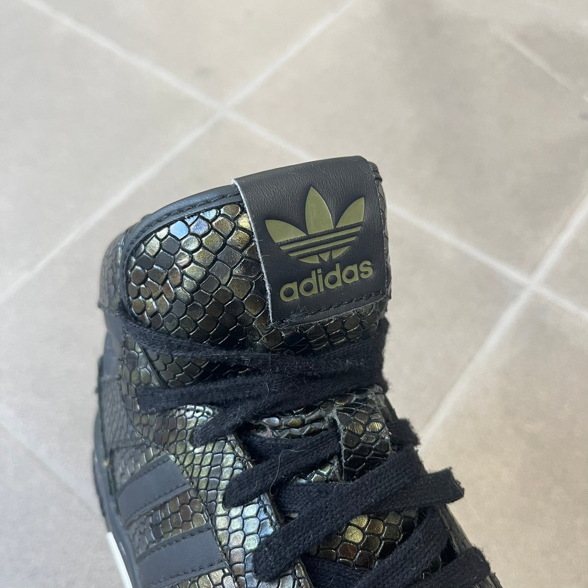 Adidas 2016 M Attitude Revive Snakeskin Sneakers are dark green sneakers covered in a shiny snakeskin-textured material, featuring black side stripes and a dark green Adidas Trefoil logo on the tongue. The sole is white with black accents.