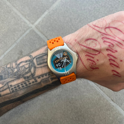 Adidas 90s Streetball 2 Analog Watch features a silver-tone metal case with a blue dial. The face includes an Adidas logo, orange “Streetball 2” branding, and white “90:78” graphics. It is equipped with a Japan movement and water-resistant construction. The watch is finished with orange rubber straps, which are a replacement/non-original strap design.
