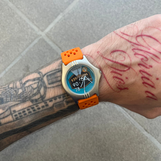 Adidas 90s Streetball 2 Analog Watch features a silver-tone metal case with a blue dial. The face includes an Adidas logo, orange “Streetball 2” branding, and white “90:78” graphics. It is equipped with a Japan movement and water-resistant construction. The watch is finished with orange rubber straps, which are a replacement/non-original strap design.
