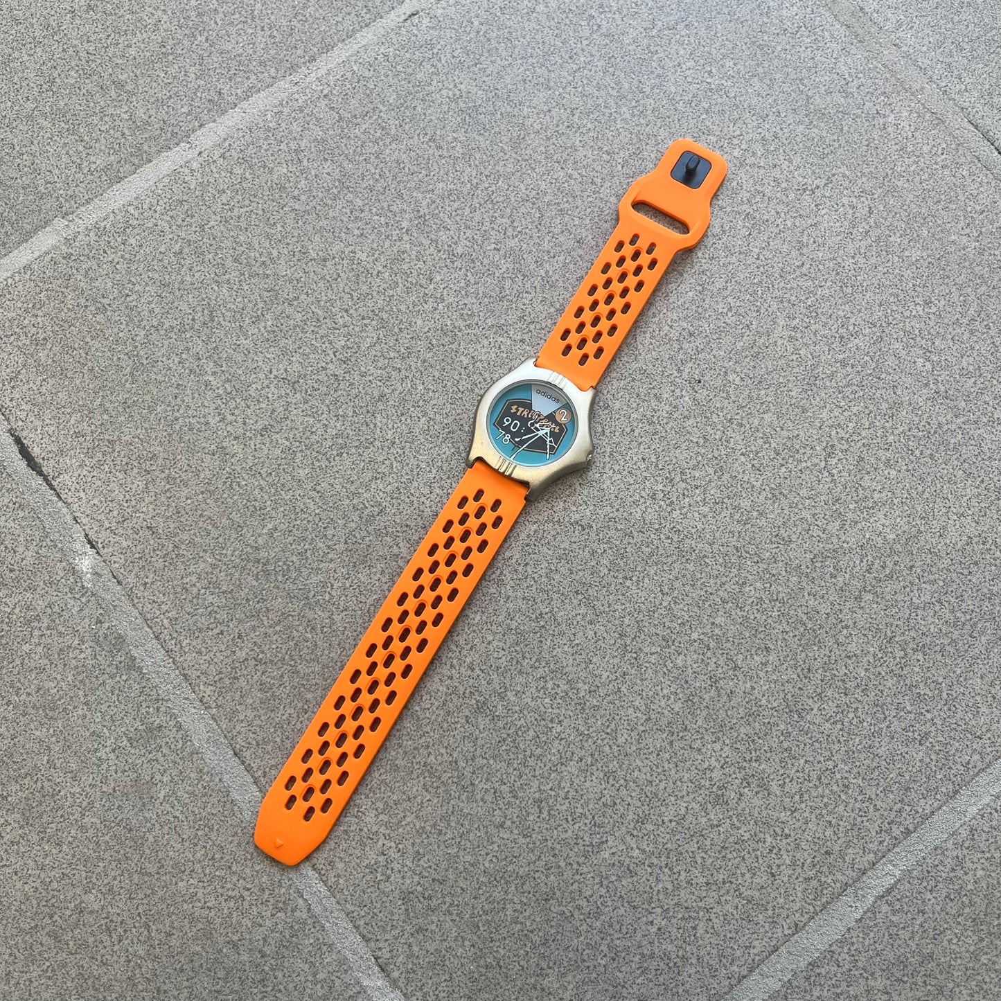 Adidas 90s Streetball 2 Analog Watch features a silver-tone metal case with a blue dial. The face includes an Adidas logo, orange “Streetball 2” branding, and white “90:78” graphics. It is equipped with a Japan movement and water-resistant construction. The watch is finished with orange rubber straps, which are a replacement/non-original strap design.
