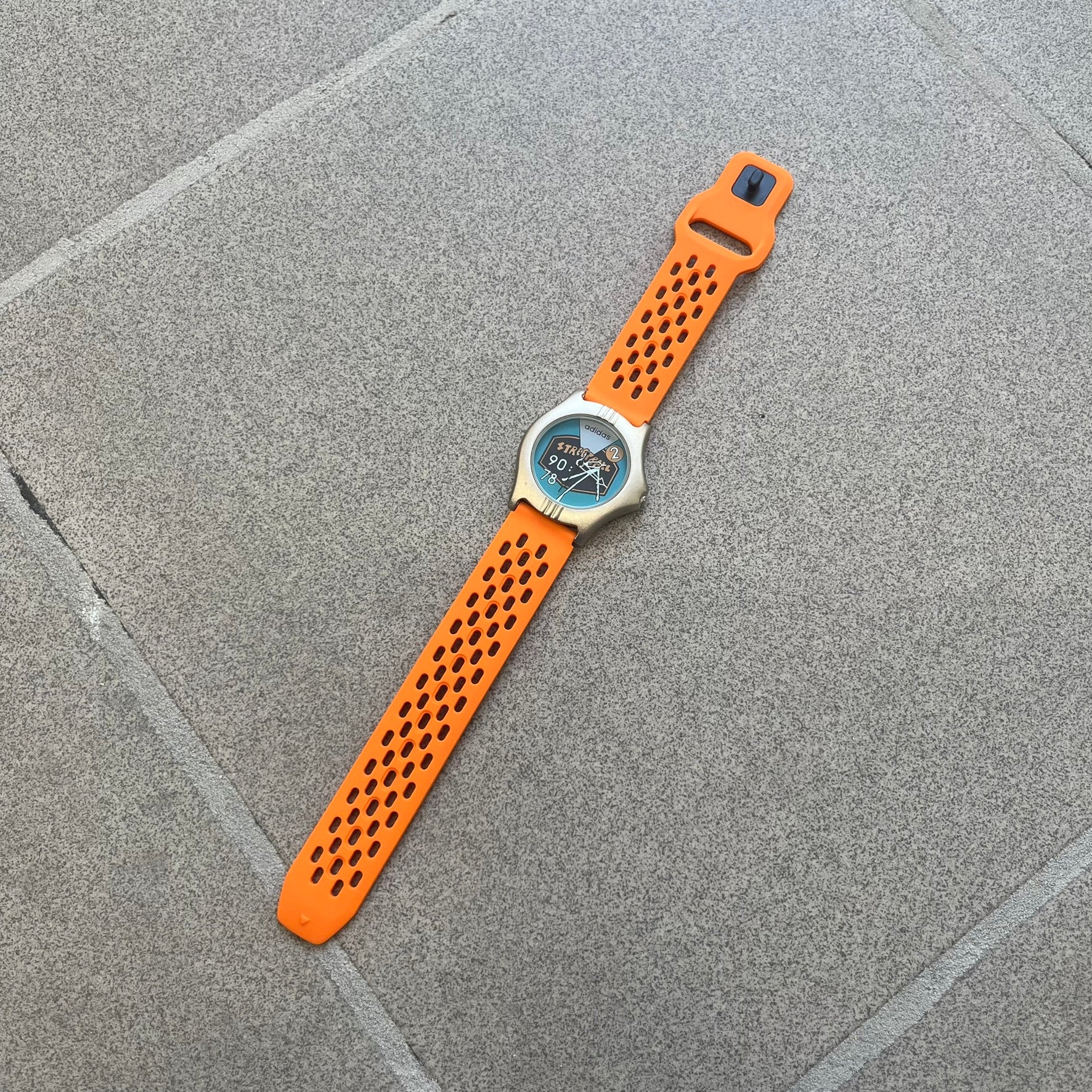 Adidas 90s Streetball 2 Analog Watch features a silver-tone metal case with a blue dial. The face includes an Adidas logo, orange “Streetball 2” branding, and white “90:78” graphics. It is equipped with a Japan movement and water-resistant construction. The watch is finished with orange rubber straps, which are a replacement/non-original strap design.
