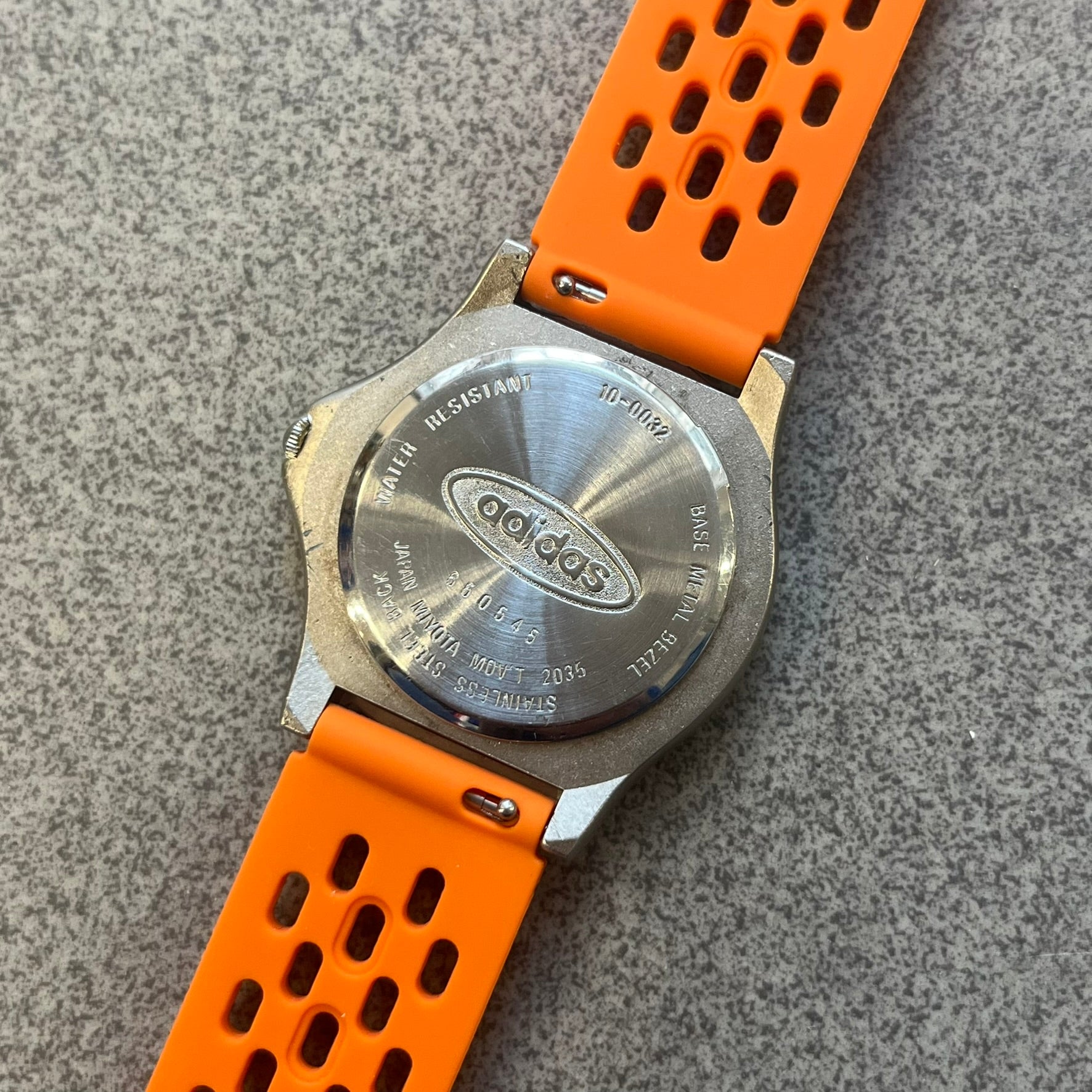 Adidas 90s Streetball 2 Analog Watch features a silver-tone metal case with a blue dial. The face includes an Adidas logo, orange “Streetball 2” branding, and white “90:78” graphics. It is equipped with a Japan movement and water-resistant construction. The watch is finished with orange rubber straps, which are a replacement/non-original strap design.
