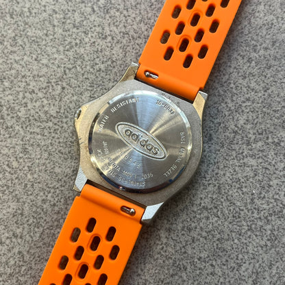 Adidas 90s Streetball 2 Analog Watch features a silver-tone metal case with a blue dial. The face includes an Adidas logo, orange “Streetball 2” branding, and white “90:78” graphics. It is equipped with a Japan movement and water-resistant construction. The watch is finished with orange rubber straps, which are a replacement/non-original strap design.
