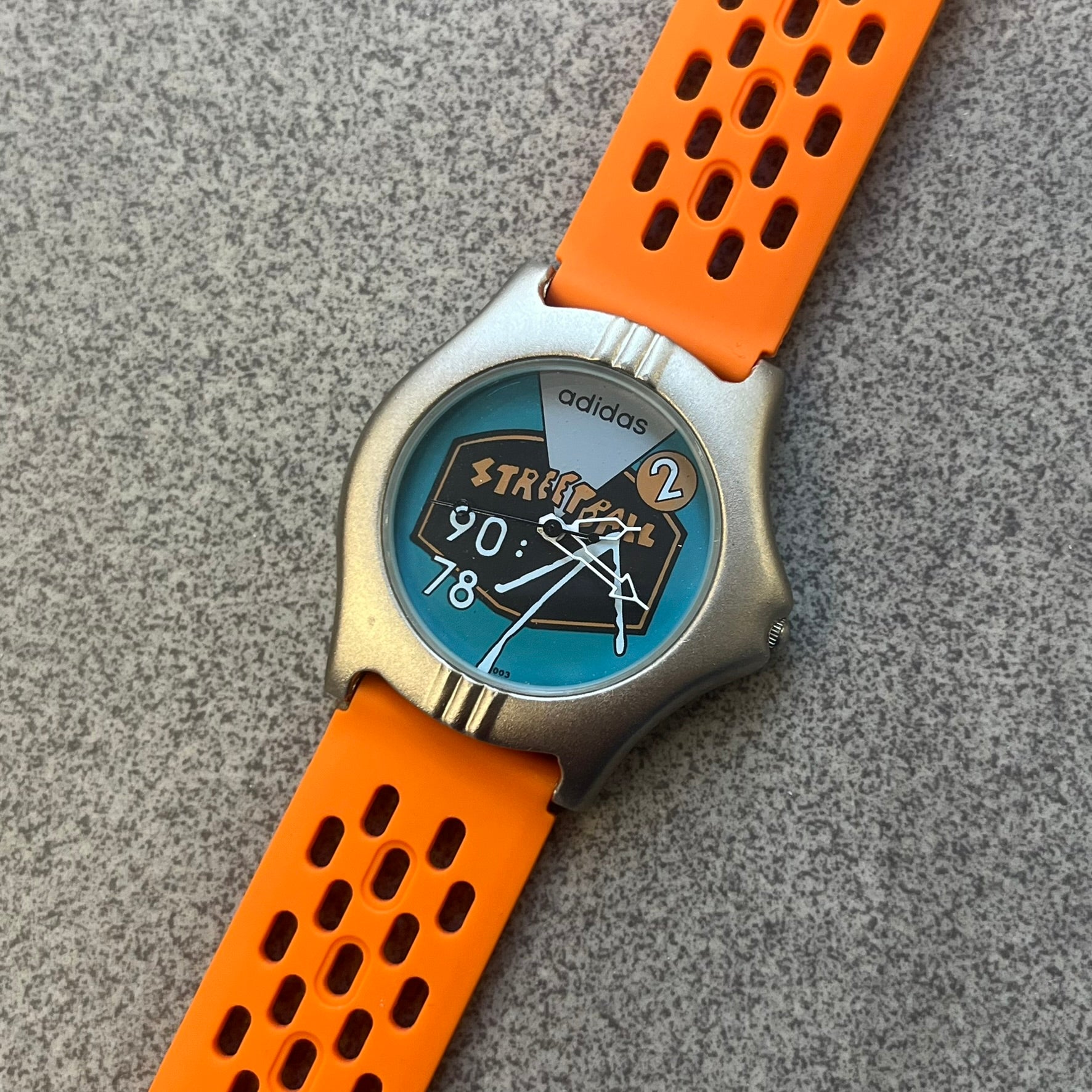 Adidas 90s Streetball 2 Analog Watch features a silver-tone metal case with a blue dial. The face includes an Adidas logo, orange “Streetball 2” branding, and white “90:78” graphics. It is equipped with a Japan movement and water-resistant construction. The watch is finished with orange rubber straps, which are a replacement/non-original strap design.
