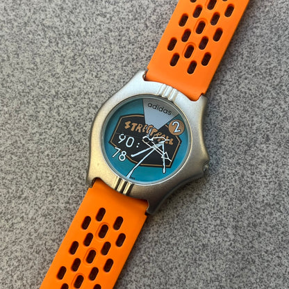 Adidas 90s Streetball 2 Analog Watch features a silver-tone metal case with a blue dial. The face includes an Adidas logo, orange “Streetball 2” branding, and white “90:78” graphics. It is equipped with a Japan movement and water-resistant construction. The watch is finished with orange rubber straps, which are a replacement/non-original strap design.
