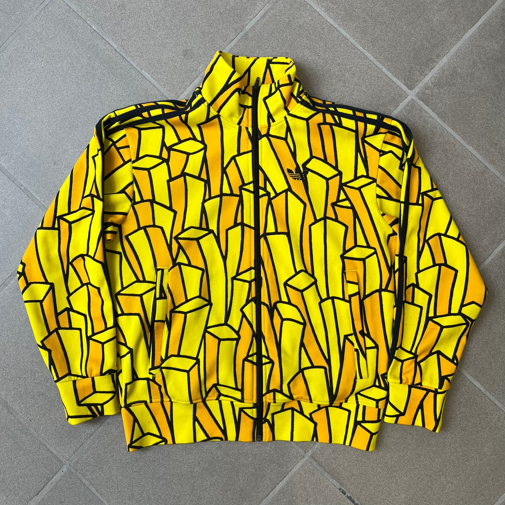 Adidas Originals x Jeremy Scott 2010 French Fry Track Jacket features an all-over yellow and black cartoon French fries print. The jacket is detailed with black Three Stripes along the sleeves, a small embroidered Trefoil logo on the chest, and a large black Trefoil with Adidas text printed across the back.