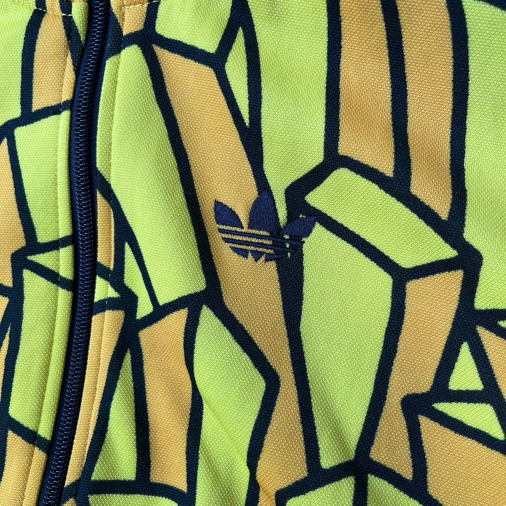 Adidas Originals x Jeremy Scott 2010 French Fry Track Jacket features an all-over yellow and black cartoon French fries print. The jacket is detailed with black Three Stripes along the sleeves, a small embroidered Trefoil logo on the chest, and a large black Trefoil with Adidas text printed across the back.