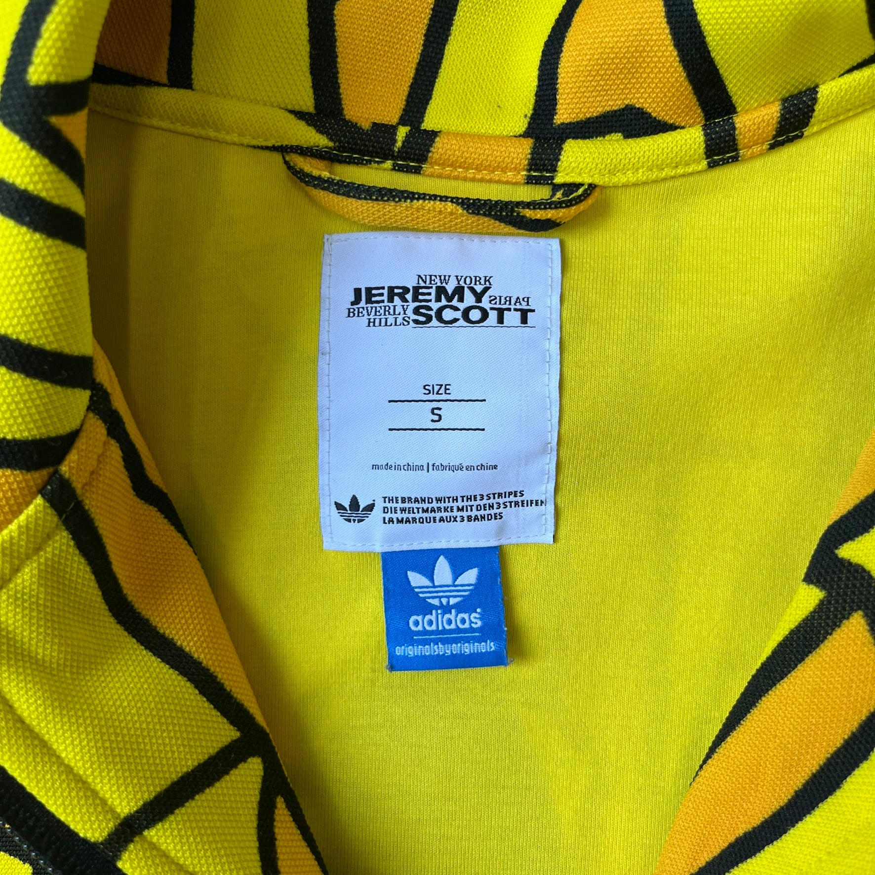 Adidas Originals x Jeremy Scott 2010 French Fry Track Jacket features an all-over yellow and black cartoon French fries print. The jacket is detailed with black Three Stripes along the sleeves, a small embroidered Trefoil logo on the chest, and a large black Trefoil with Adidas text printed across the back.