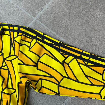 Adidas Originals x Jeremy Scott 2010 French Fry Track Jacket features an all-over yellow and black cartoon French fries print. The jacket is detailed with black Three Stripes along the sleeves, a small embroidered Trefoil logo on the chest, and a large black Trefoil with Adidas text printed across the back.