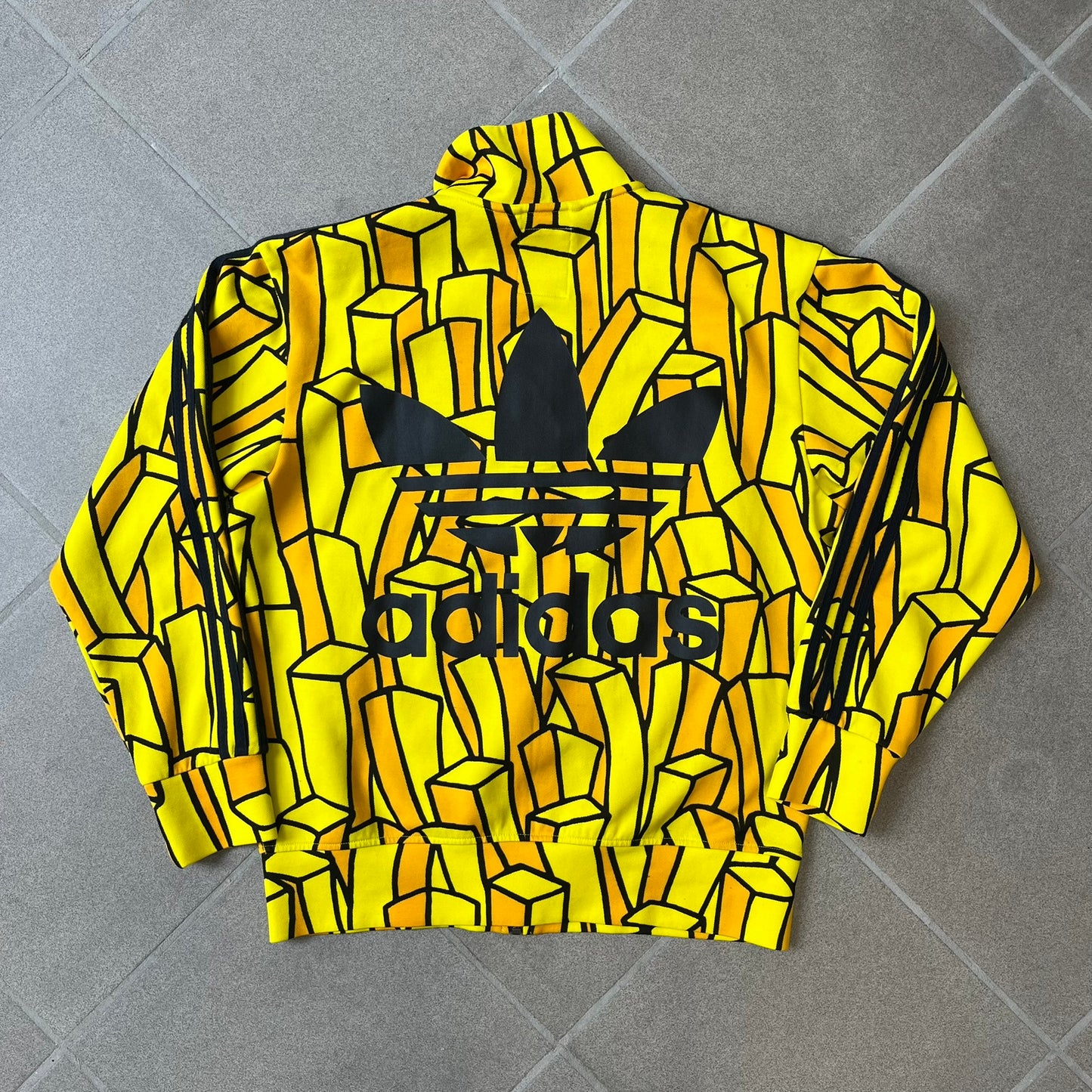 Adidas Originals x Jeremy Scott 2010 French Fry Track Jacket features an all-over yellow and black cartoon French fries print. The jacket is detailed with black Three Stripes along the sleeves, a small embroidered Trefoil logo on the chest, and a large black Trefoil with Adidas text printed across the back.