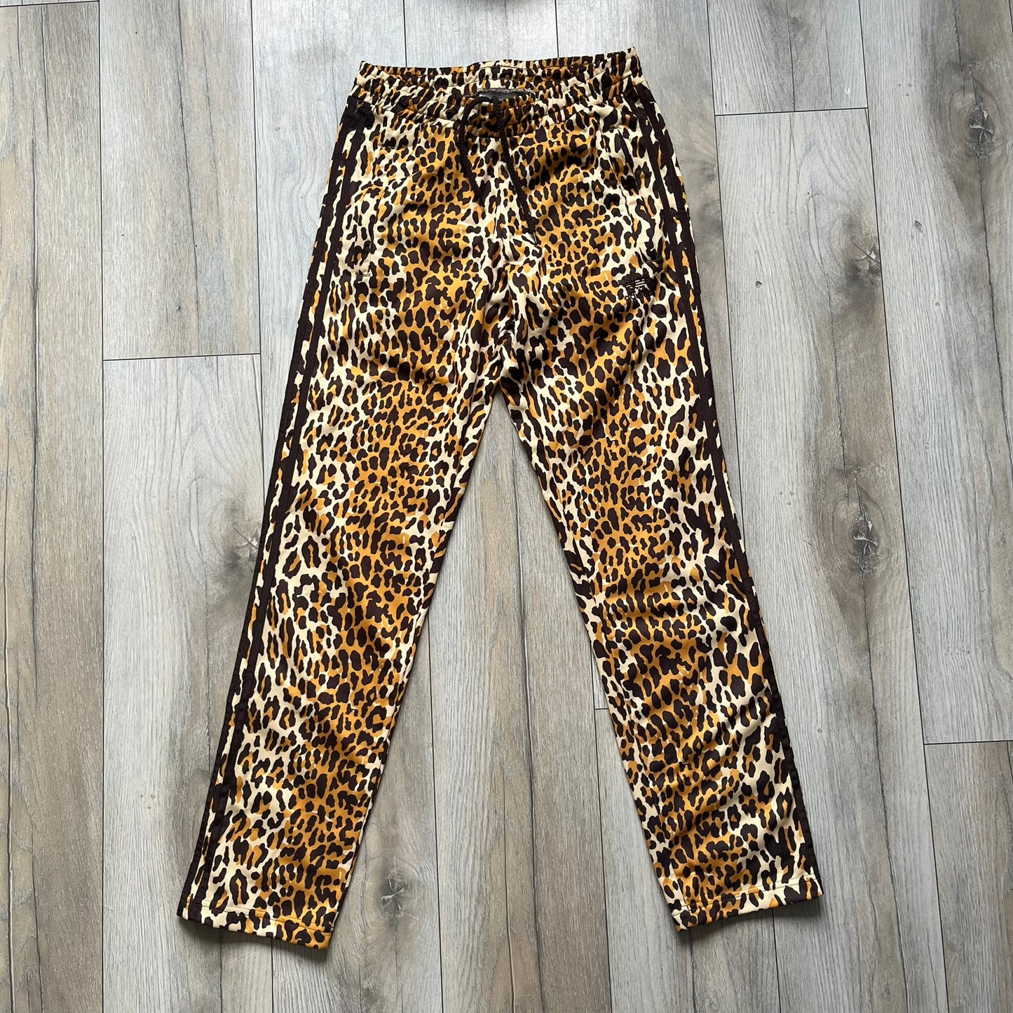 Adidas x Jeremy Scott 2015 Leopard Print Track Pants feature an all-over orange, brown, and white leopard print pattern throughout the garment. The design includes brown Adidas three-stripe detailing down both sides, zippered pockets, brown drawstrings, and an embroidered Adidas Trefoil logo. This pair was released as part of the Adidas Originals by Jeremy Scott collaboration.