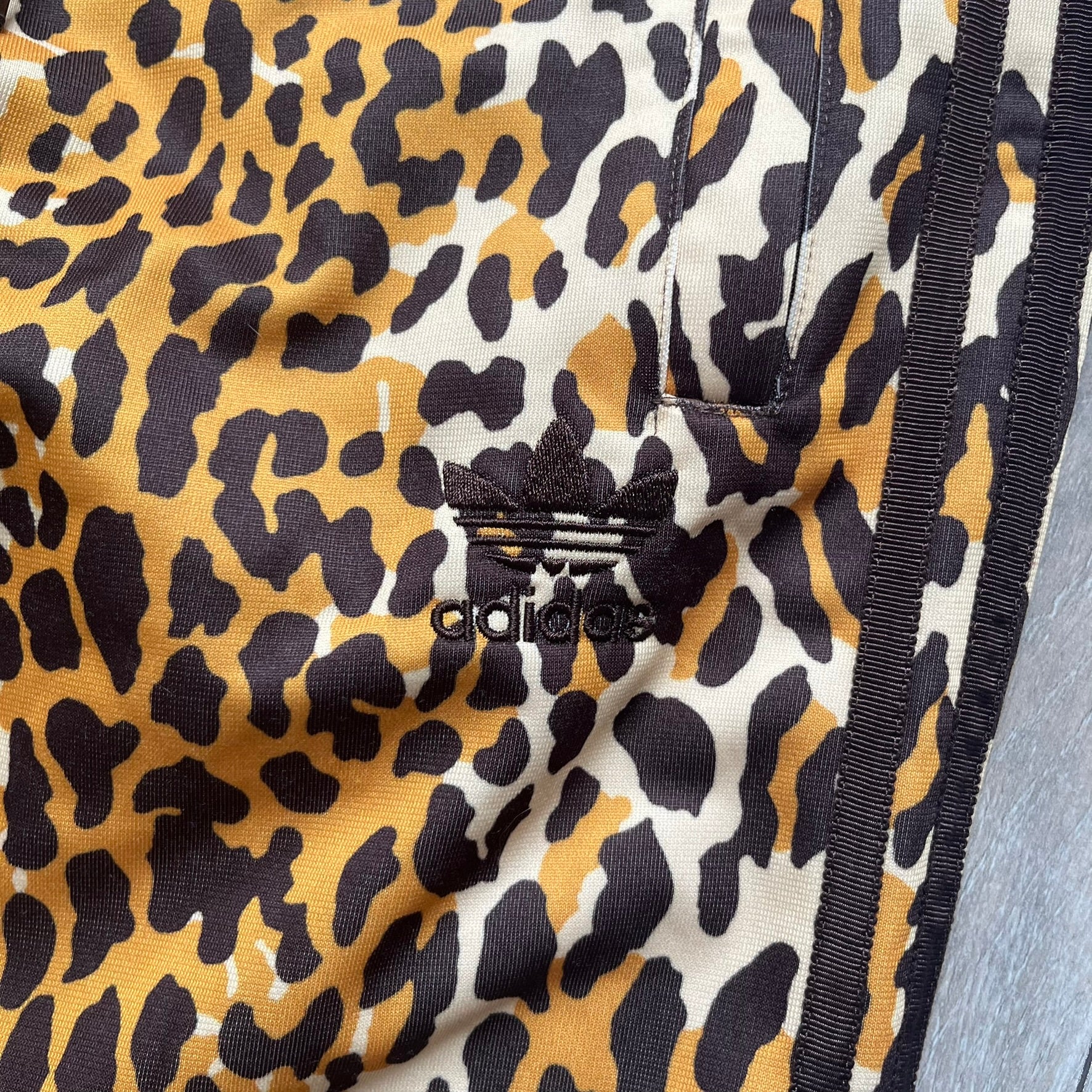 Adidas x Jeremy Scott 2015 Leopard Print Track Pants feature an all-over orange, brown, and white leopard print pattern throughout the garment. The design includes brown Adidas three-stripe detailing down both sides, zippered pockets, brown drawstrings, and an embroidered Adidas Trefoil logo. This pair was released as part of the Adidas Originals by Jeremy Scott collaboration.