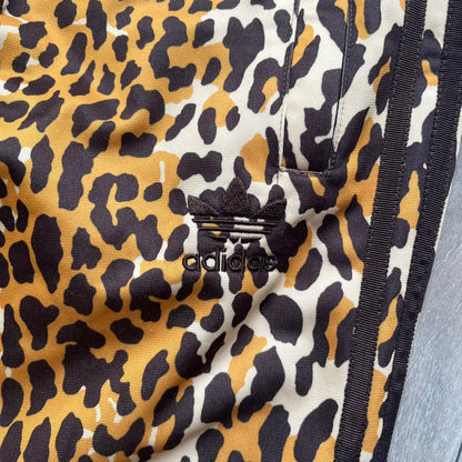 Adidas x Jeremy Scott 2015 Leopard Print Track Pants feature an all-over orange, brown, and white leopard print pattern throughout the garment. The design includes brown Adidas three-stripe detailing down both sides, zippered pockets, brown drawstrings, and an embroidered Adidas Trefoil logo. This pair was released as part of the Adidas Originals by Jeremy Scott collaboration.