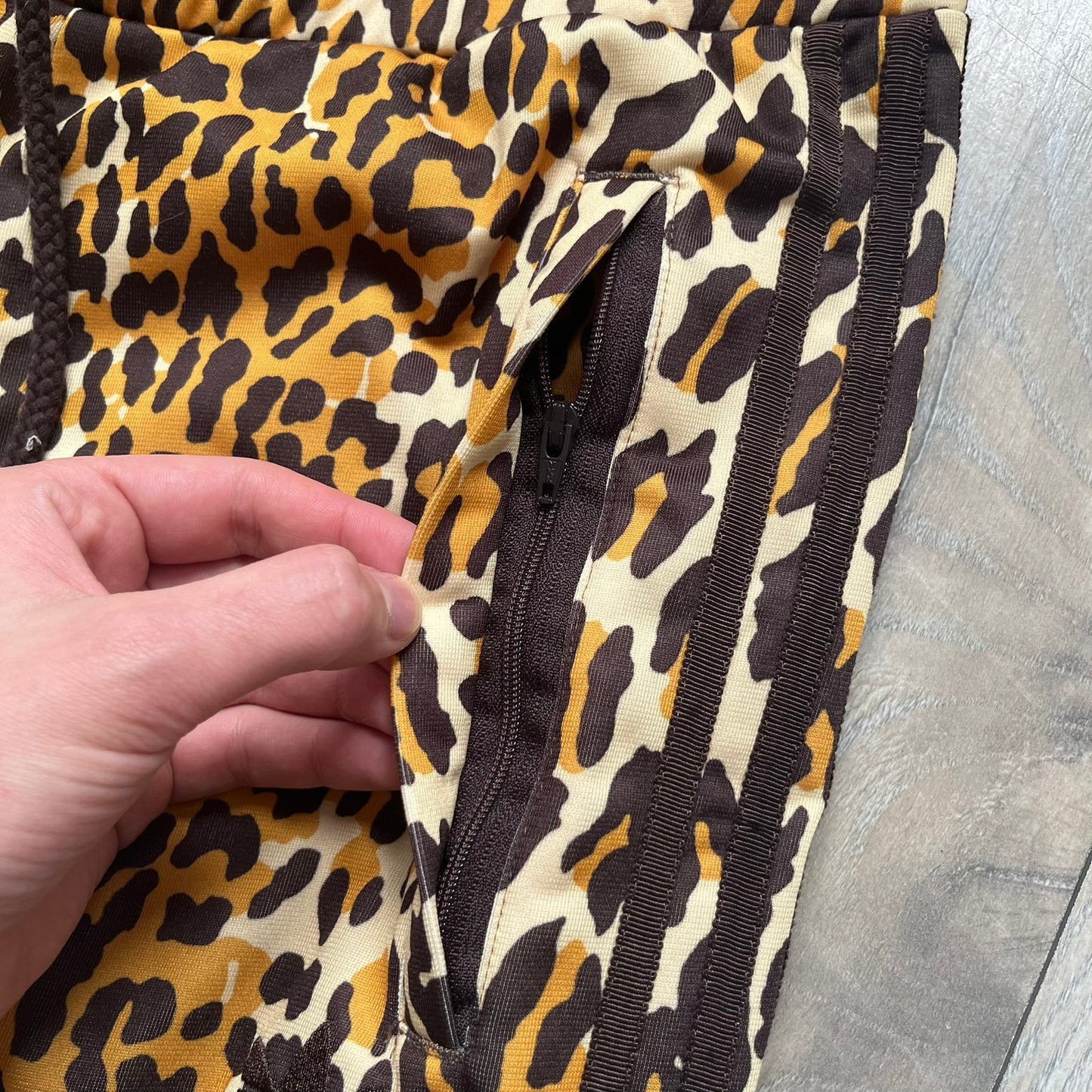 Adidas x Jeremy Scott 2015 Leopard Print Track Pants feature an all-over orange, brown, and white leopard print pattern throughout the garment. The design includes brown Adidas three-stripe detailing down both sides, zippered pockets, brown drawstrings, and an embroidered Adidas Trefoil logo. This pair was released as part of the Adidas Originals by Jeremy Scott collaboration.
