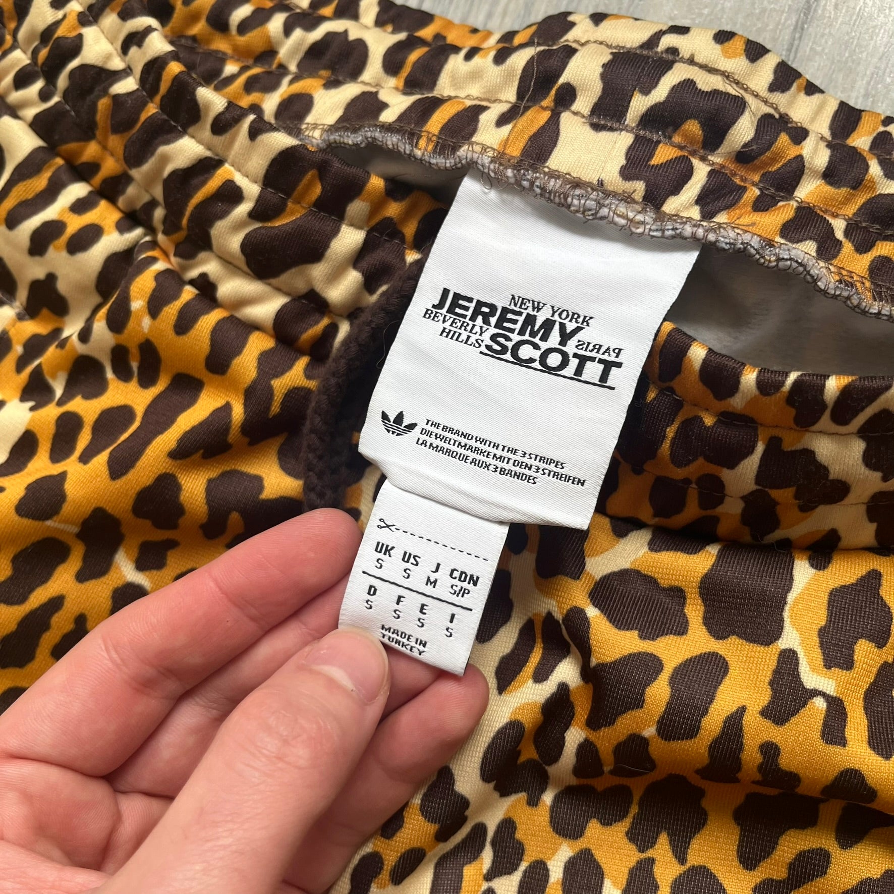 Adidas x Jeremy Scott 2015 Leopard Print Track Pants feature an all-over orange, brown, and white leopard print pattern throughout the garment. The design includes brown Adidas three-stripe detailing down both sides, zippered pockets, brown drawstrings, and an embroidered Adidas Trefoil logo. This pair was released as part of the Adidas Originals by Jeremy Scott collaboration.