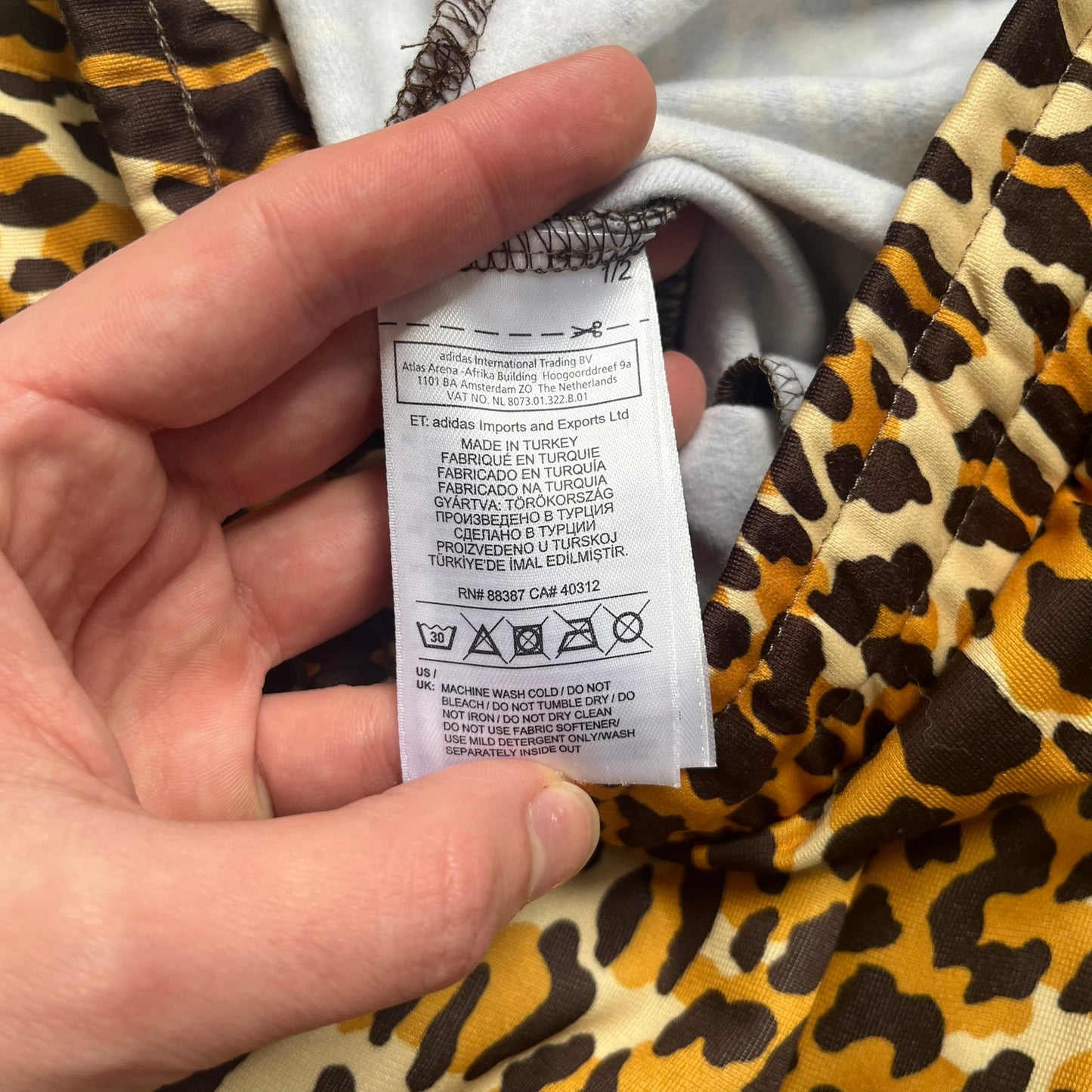Adidas x Jeremy Scott 2015 Leopard Print Track Pants feature an all-over orange, brown, and white leopard print pattern throughout the garment. The design includes brown Adidas three-stripe detailing down both sides, zippered pockets, brown drawstrings, and an embroidered Adidas Trefoil logo. This pair was released as part of the Adidas Originals by Jeremy Scott collaboration.