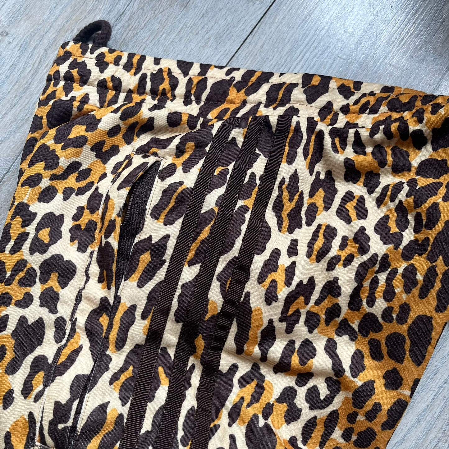 Adidas x Jeremy Scott 2015 Leopard Print Track Pants feature an all-over orange, brown, and white leopard print pattern throughout the garment. The design includes brown Adidas three-stripe detailing down both sides, zippered pockets, brown drawstrings, and an embroidered Adidas Trefoil logo. This pair was released as part of the Adidas Originals by Jeremy Scott collaboration.