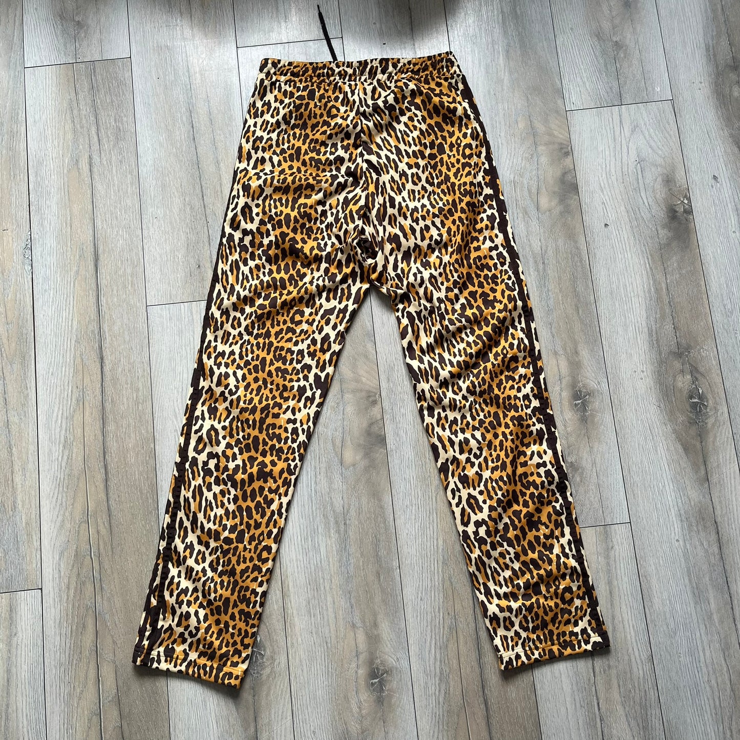 Adidas x Jeremy Scott 2015 Leopard Print Track Pants feature an all-over orange, brown, and white leopard print pattern throughout the garment. The design includes brown Adidas three-stripe detailing down both sides, zippered pockets, brown drawstrings, and an embroidered Adidas Trefoil logo. This pair was released as part of the Adidas Originals by Jeremy Scott collaboration.