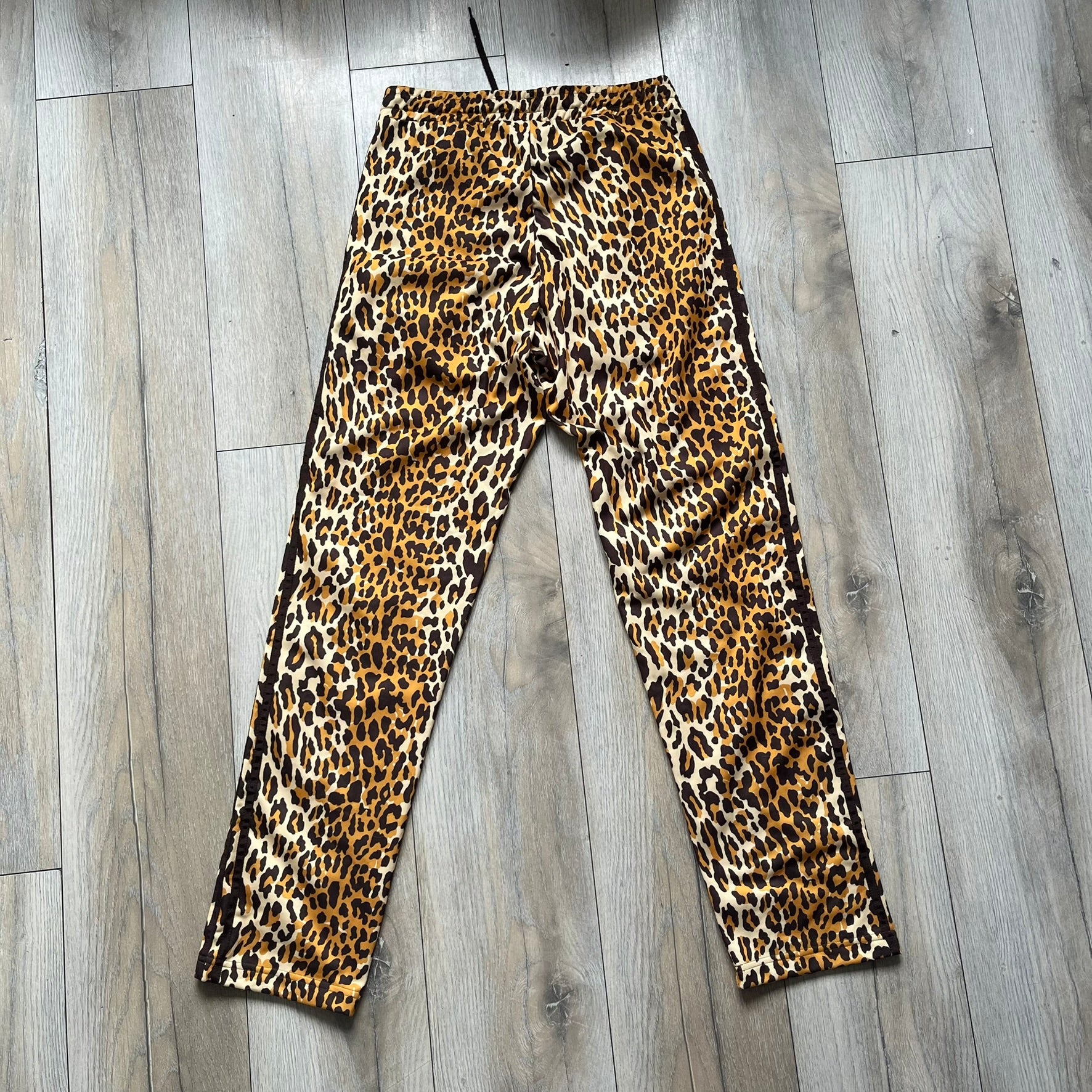 Adidas x Jeremy Scott 2015 Leopard Print Track Pants feature an all-over orange, brown, and white leopard print pattern throughout the garment. The design includes brown Adidas three-stripe detailing down both sides, zippered pockets, brown drawstrings, and an embroidered Adidas Trefoil logo. This pair was released as part of the Adidas Originals by Jeremy Scott collaboration.