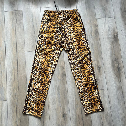 Adidas x Jeremy Scott 2015 Leopard Print Track Pants feature an all-over orange, brown, and white leopard print pattern throughout the garment. The design includes brown Adidas three-stripe detailing down both sides, zippered pockets, brown drawstrings, and an embroidered Adidas Trefoil logo. This pair was released as part of the Adidas Originals by Jeremy Scott collaboration.