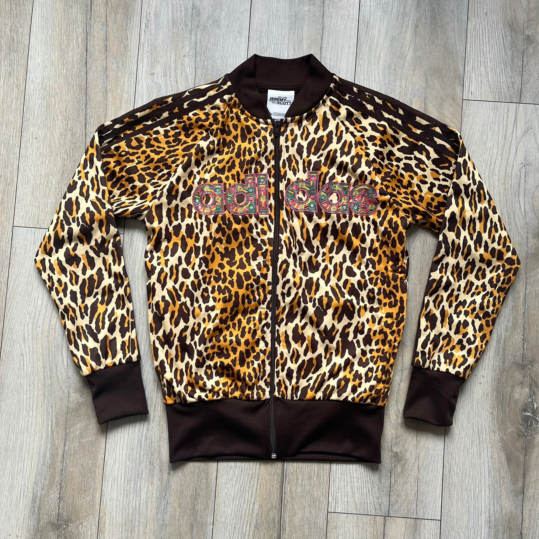 Adidas x Jeremy Scott 2015 Leopard Sequin Track Jacket features an all-over orange, brown, and white leopard print pattern with brown hems throughout. The jacket includes brown Adidas three-stripe detailing on the shoulders and sleeves, zippered front pockets, and multicolor embroidered sequin “adi” and “das” chest branding in Jeremy Scott styling. The back is finished with a large embroidered sequin Adidas Trefoil logo, and the piece comes with original paper tag.