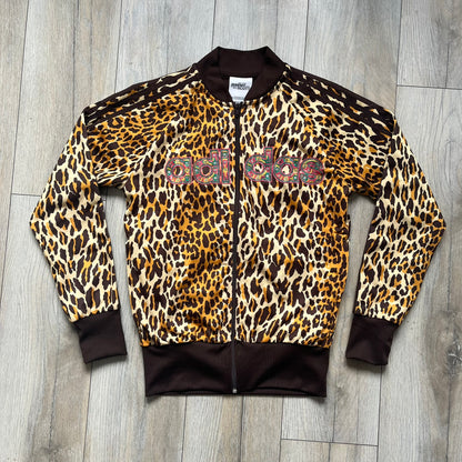 Adidas x Jeremy Scott 2015 Leopard Sequin Track Jacket features an all-over orange, brown, and white leopard print pattern with brown hems throughout. The jacket includes brown Adidas three-stripe detailing on the shoulders and sleeves, zippered front pockets, and multicolor embroidered sequin “adi” and “das” chest branding in Jeremy Scott styling. The back is finished with a large embroidered sequin Adidas Trefoil logo, and the piece comes with original paper tag.