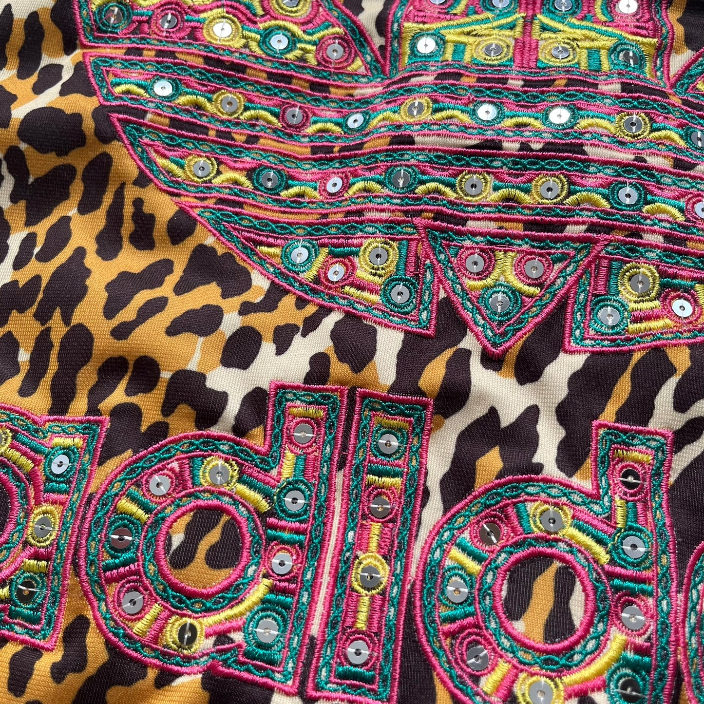 Adidas x Jeremy Scott 2015 Leopard Sequin Track Jacket features an all-over orange, brown, and white leopard print pattern with brown hems throughout. The jacket includes brown Adidas three-stripe detailing on the shoulders and sleeves, zippered front pockets, and multicolor embroidered sequin “adi” and “das” chest branding in Jeremy Scott styling. The back is finished with a large embroidered sequin Adidas Trefoil logo, and the piece comes with original paper tag.