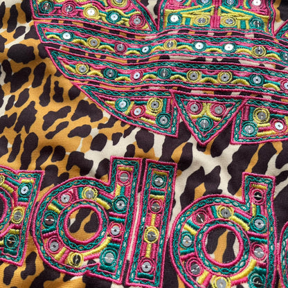 Adidas x Jeremy Scott 2015 Leopard Sequin Track Jacket features an all-over orange, brown, and white leopard print pattern with brown hems throughout. The jacket includes brown Adidas three-stripe detailing on the shoulders and sleeves, zippered front pockets, and multicolor embroidered sequin “adi” and “das” chest branding in Jeremy Scott styling. The back is finished with a large embroidered sequin Adidas Trefoil logo, and the piece comes with original paper tag.