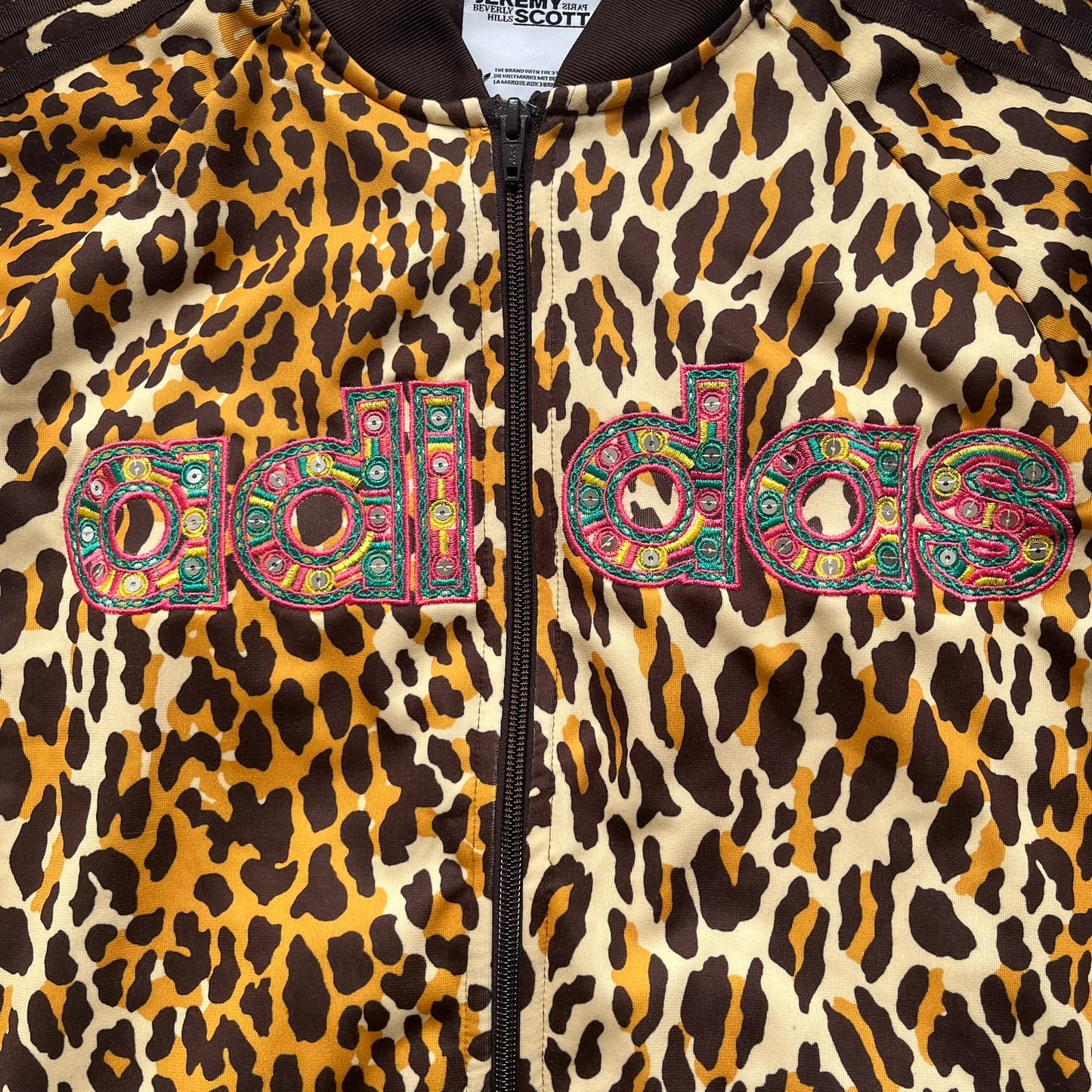 Adidas x Jeremy Scott 2015 Leopard Sequin Track Jacket features an all-over orange, brown, and white leopard print pattern with brown hems throughout. The jacket includes brown Adidas three-stripe detailing on the shoulders and sleeves, zippered front pockets, and multicolor embroidered sequin “adi” and “das” chest branding in Jeremy Scott styling. The back is finished with a large embroidered sequin Adidas Trefoil logo, and the piece comes with original paper tag.