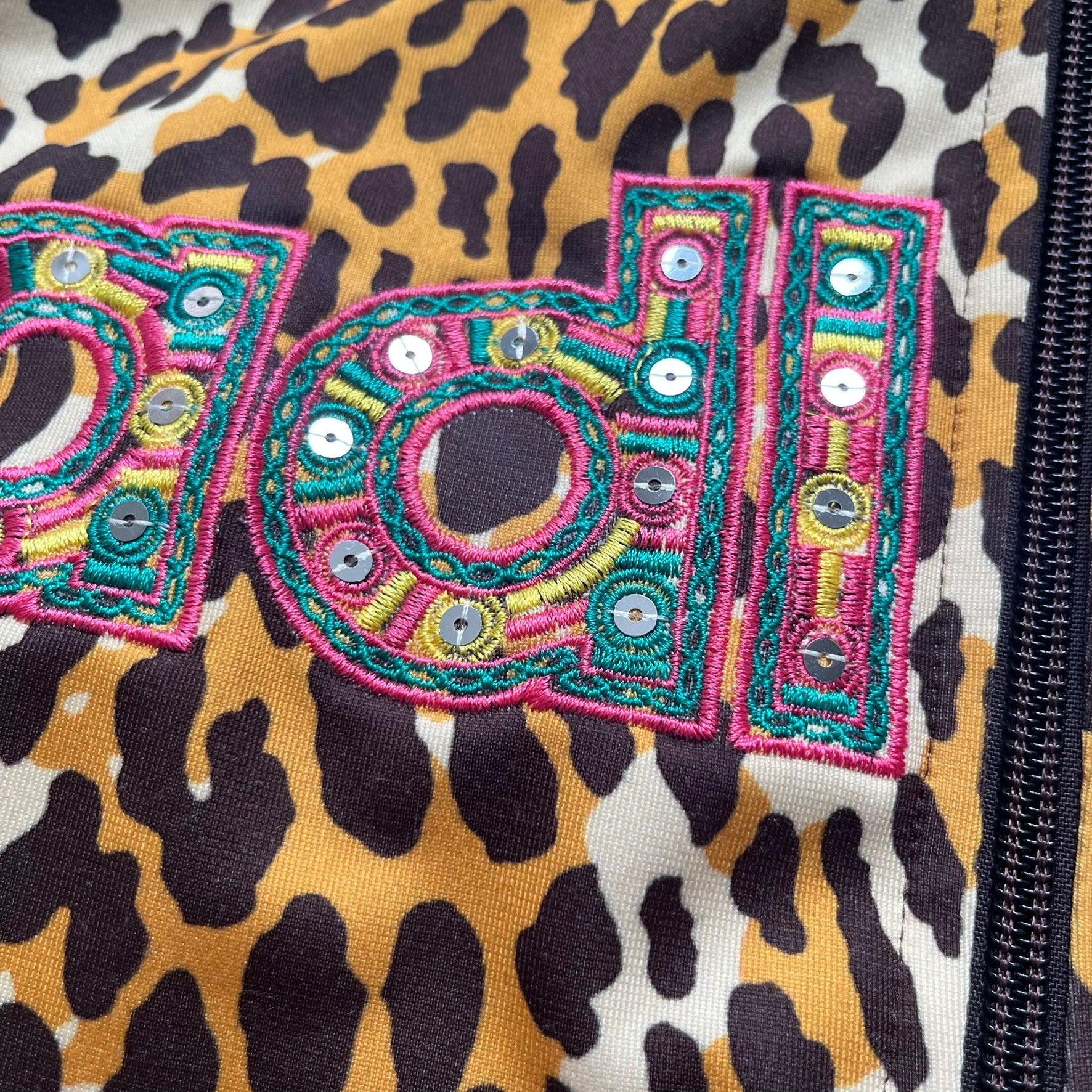 Adidas x Jeremy Scott 2015 Leopard Sequin Track Jacket features an all-over orange, brown, and white leopard print pattern with brown hems throughout. The jacket includes brown Adidas three-stripe detailing on the shoulders and sleeves, zippered front pockets, and multicolor embroidered sequin “adi” and “das” chest branding in Jeremy Scott styling. The back is finished with a large embroidered sequin Adidas Trefoil logo, and the piece comes with original paper tag.