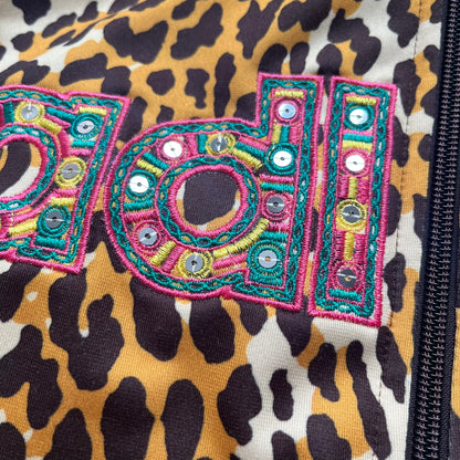 Adidas x Jeremy Scott 2015 Leopard Sequin Track Jacket features an all-over orange, brown, and white leopard print pattern with brown hems throughout. The jacket includes brown Adidas three-stripe detailing on the shoulders and sleeves, zippered front pockets, and multicolor embroidered sequin “adi” and “das” chest branding in Jeremy Scott styling. The back is finished with a large embroidered sequin Adidas Trefoil logo, and the piece comes with original paper tag.