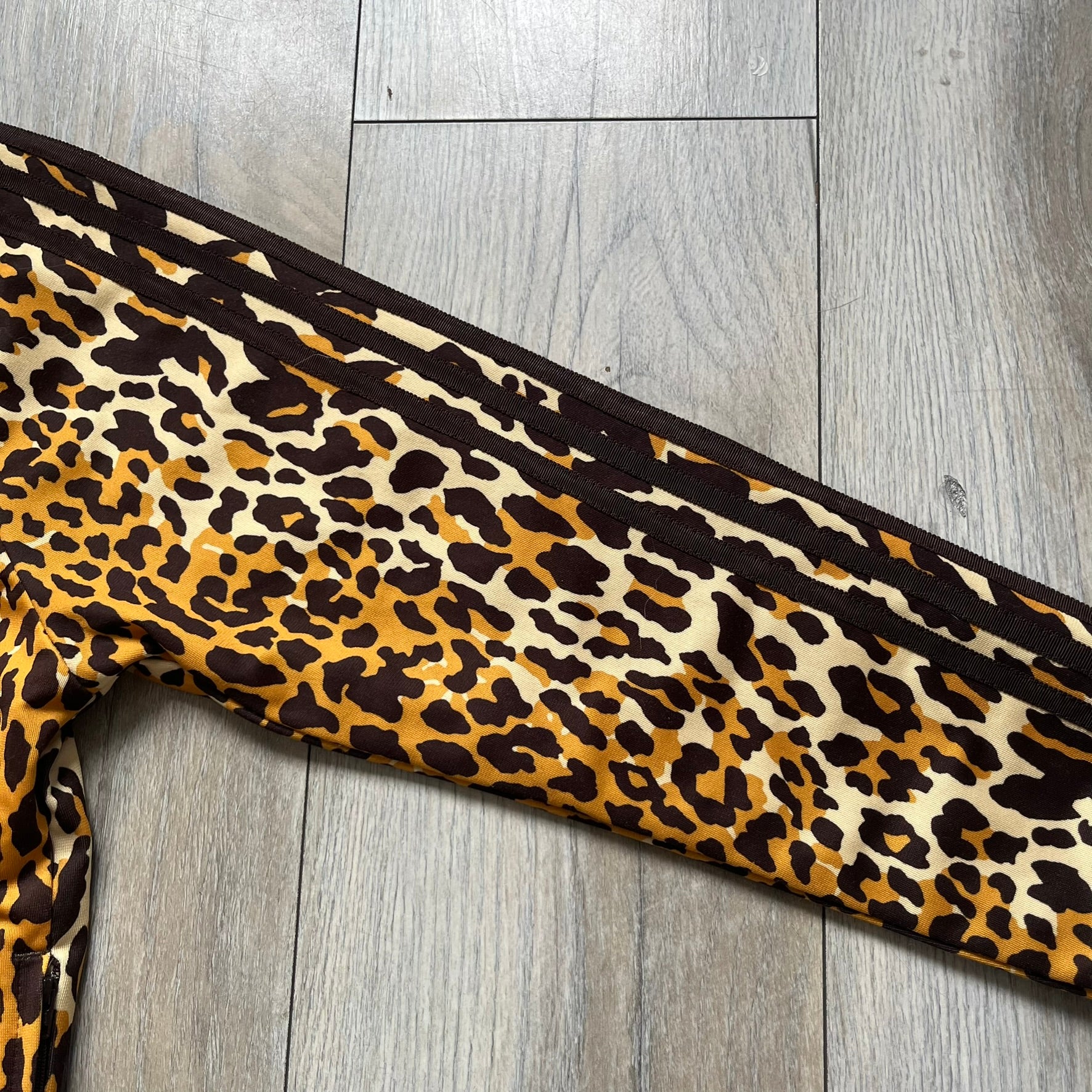 Adidas x Jeremy Scott 2015 Leopard Sequin Track Jacket features an all-over orange, brown, and white leopard print pattern with brown hems throughout. The jacket includes brown Adidas three-stripe detailing on the shoulders and sleeves, zippered front pockets, and multicolor embroidered sequin “adi” and “das” chest branding in Jeremy Scott styling. The back is finished with a large embroidered sequin Adidas Trefoil logo, and the piece comes with original paper tag.
