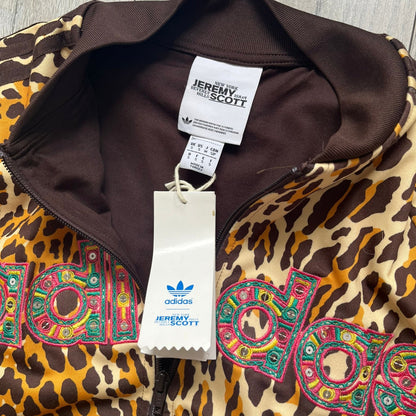 Adidas x Jeremy Scott 2015 Leopard Sequin Track Jacket features an all-over orange, brown, and white leopard print pattern with brown hems throughout. The jacket includes brown Adidas three-stripe detailing on the shoulders and sleeves, zippered front pockets, and multicolor embroidered sequin “adi” and “das” chest branding in Jeremy Scott styling. The back is finished with a large embroidered sequin Adidas Trefoil logo, and the piece comes with original paper tag.