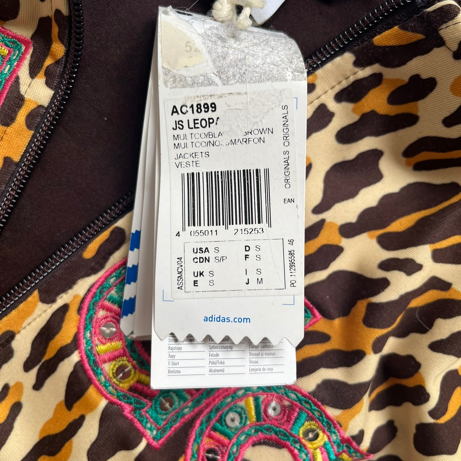 Adidas x Jeremy Scott 2015 Leopard Sequin Track Jacket features an all-over orange, brown, and white leopard print pattern with brown hems throughout. The jacket includes brown Adidas three-stripe detailing on the shoulders and sleeves, zippered front pockets, and multicolor embroidered sequin “adi” and “das” chest branding in Jeremy Scott styling. The back is finished with a large embroidered sequin Adidas Trefoil logo, and the piece comes with original paper tag.