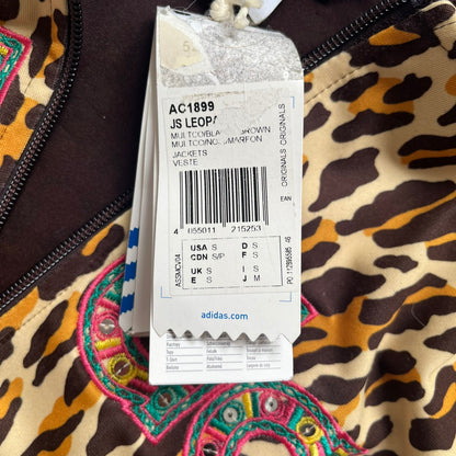 Adidas x Jeremy Scott 2015 Leopard Sequin Track Jacket features an all-over orange, brown, and white leopard print pattern with brown hems throughout. The jacket includes brown Adidas three-stripe detailing on the shoulders and sleeves, zippered front pockets, and multicolor embroidered sequin “adi” and “das” chest branding in Jeremy Scott styling. The back is finished with a large embroidered sequin Adidas Trefoil logo, and the piece comes with original paper tag.