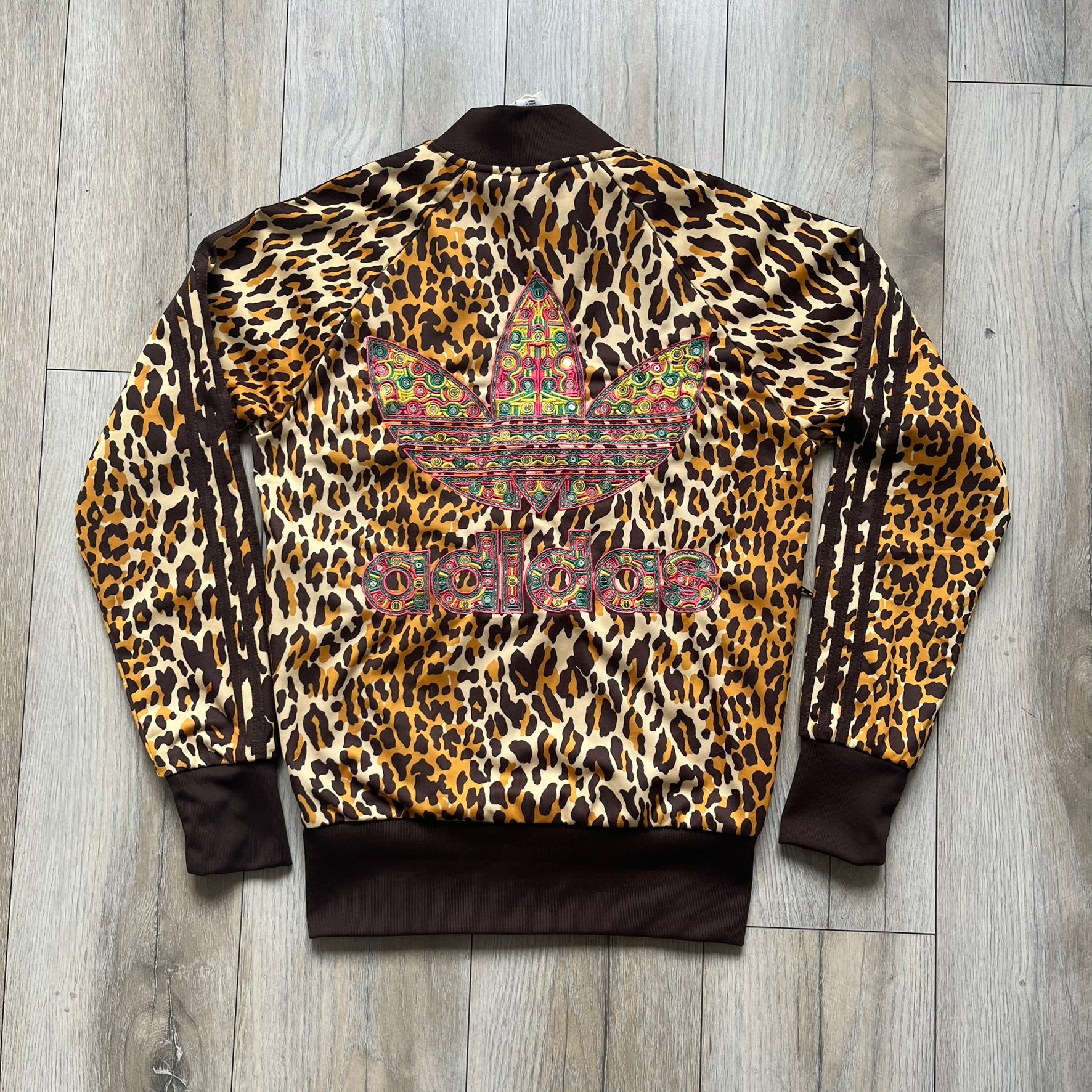 Adidas x Jeremy Scott 2015 Leopard Sequin Track Jacket features an all-over orange, brown, and white leopard print pattern with brown hems throughout. The jacket includes brown Adidas three-stripe detailing on the shoulders and sleeves, zippered front pockets, and multicolor embroidered sequin “adi” and “das” chest branding in Jeremy Scott styling. The back is finished with a large embroidered sequin Adidas Trefoil logo, and the piece comes with original paper tag.