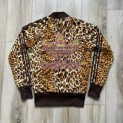 Adidas x Jeremy Scott 2015 Leopard Sequin Track Jacket features an all-over orange, brown, and white leopard print pattern with brown hems throughout. The jacket includes brown Adidas three-stripe detailing on the shoulders and sleeves, zippered front pockets, and multicolor embroidered sequin “adi” and “das” chest branding in Jeremy Scott styling. The back is finished with a large embroidered sequin Adidas Trefoil logo, and the piece comes with original paper tag.