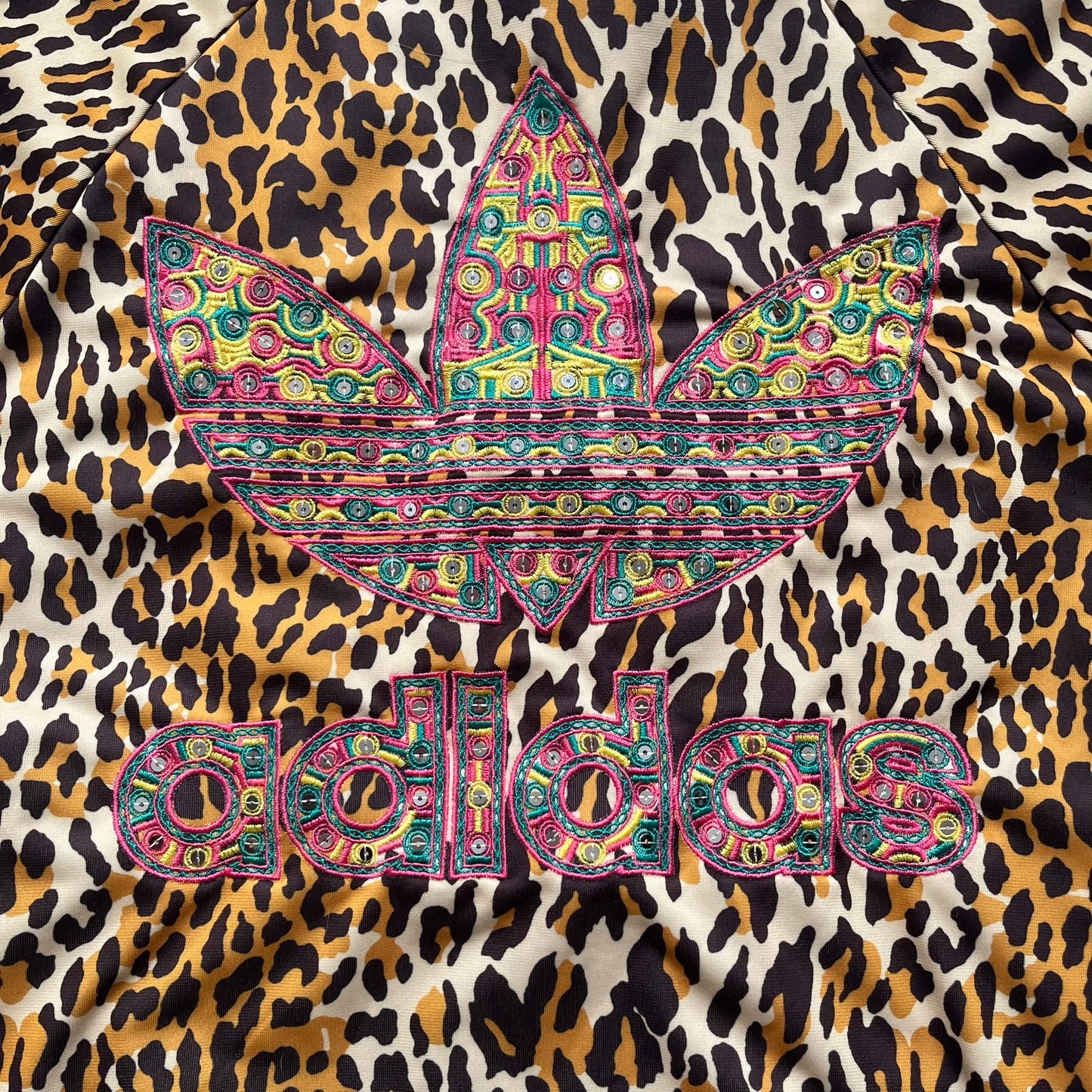 Adidas x Jeremy Scott 2015 Leopard Sequin Track Jacket features an all-over orange, brown, and white leopard print pattern with brown hems throughout. The jacket includes brown Adidas three-stripe detailing on the shoulders and sleeves, zippered front pockets, and multicolor embroidered sequin “adi” and “das” chest branding in Jeremy Scott styling. The back is finished with a large embroidered sequin Adidas Trefoil logo, and the piece comes with original paper tag.