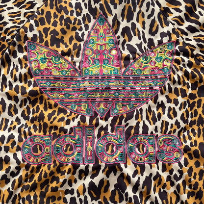Adidas x Jeremy Scott 2015 Leopard Sequin Track Jacket features an all-over orange, brown, and white leopard print pattern with brown hems throughout. The jacket includes brown Adidas three-stripe detailing on the shoulders and sleeves, zippered front pockets, and multicolor embroidered sequin “adi” and “das” chest branding in Jeremy Scott styling. The back is finished with a large embroidered sequin Adidas Trefoil logo, and the piece comes with original paper tag.