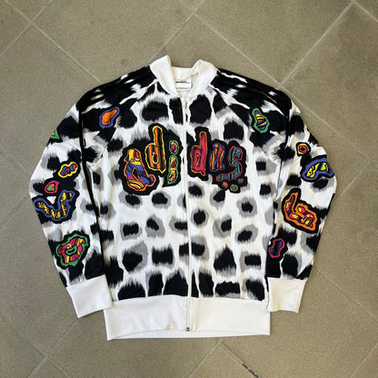 Adidas Jeremy Scott 2015 White Leopard Track Jacket is a white track jacket with a black and gray leopard print, with some of the leopard patches filled in with colorful patterns. Embroidered Adidas stripes run along the sleeves and shoulders, and colorful squiggly Adidas text appears on the chest. The back repeats the same text under a large colorful Trefoil logo, with all colored parts made of mixed patterns and colors.