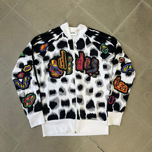 Adidas Jeremy Scott 2015 White Leopard Track Jacket is a white track jacket with a black and gray leopard print, with some of the leopard patches filled in with colorful patterns. Embroidered Adidas stripes run along the sleeves and shoulders, and colorful squiggly Adidas text appears on the chest. The back repeats the same text under a large colorful Trefoil logo, with all colored parts made of mixed patterns and colors.