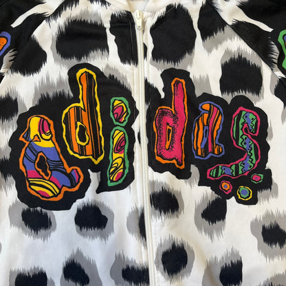 Adidas Jeremy Scott 2015 White Leopard Track Jacket is a white track jacket with a black and gray leopard print, with some of the leopard patches filled in with colorful patterns. Embroidered Adidas stripes run along the sleeves and shoulders, and colorful squiggly Adidas text appears on the chest. The back repeats the same text under a large colorful Trefoil logo, with all colored parts made of mixed patterns and colors.