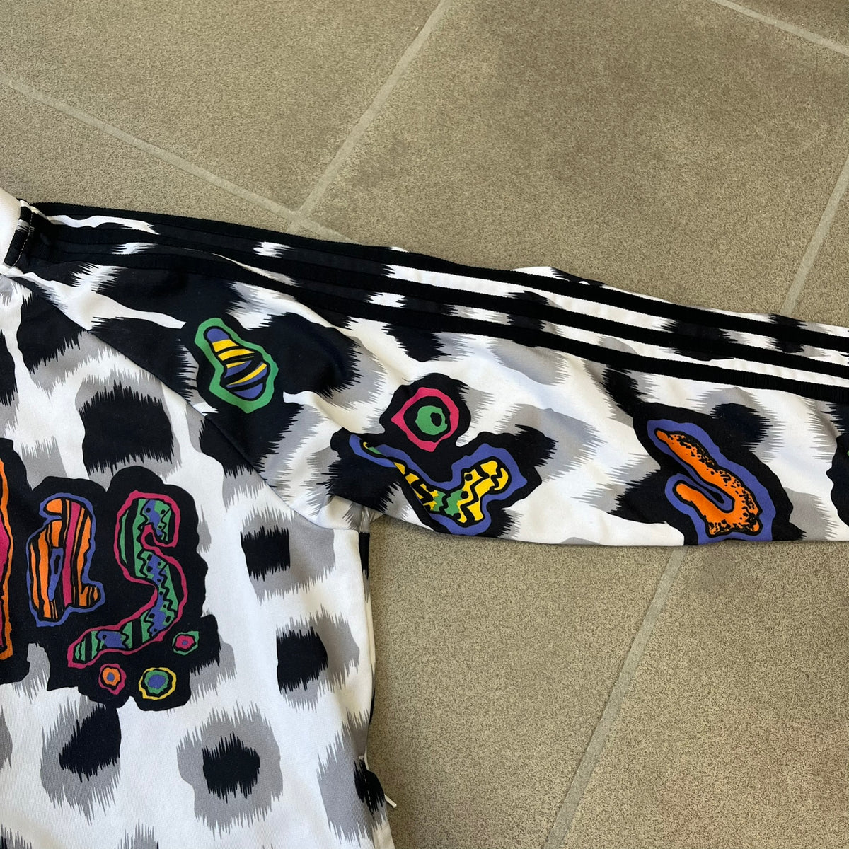 Adidas Jeremy Scott 2015 White Leopard Track Jacket is a white track jacket with a black and gray leopard print, with some of the leopard patches filled in with colorful patterns. Embroidered Adidas stripes run along the sleeves and shoulders, and colorful squiggly Adidas text appears on the chest. The back repeats the same text under a large colorful Trefoil logo, with all colored parts made of mixed patterns and colors.
