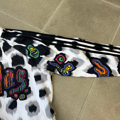Adidas Jeremy Scott 2015 White Leopard Track Jacket is a white track jacket with a black and gray leopard print, with some of the leopard patches filled in with colorful patterns. Embroidered Adidas stripes run along the sleeves and shoulders, and colorful squiggly Adidas text appears on the chest. The back repeats the same text under a large colorful Trefoil logo, with all colored parts made of mixed patterns and colors.