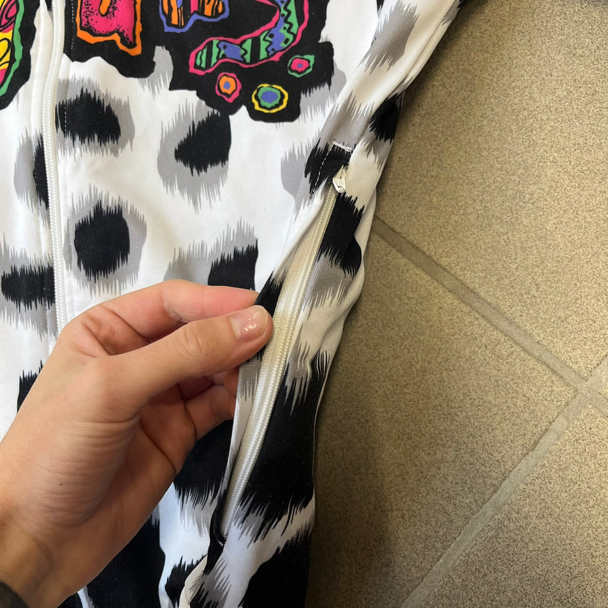 Adidas Jeremy Scott 2015 White Leopard Track Jacket is a white track jacket with a black and gray leopard print, with some of the leopard patches filled in with colorful patterns. Embroidered Adidas stripes run along the sleeves and shoulders, and colorful squiggly Adidas text appears on the chest. The back repeats the same text under a large colorful Trefoil logo, with all colored parts made of mixed patterns and colors.