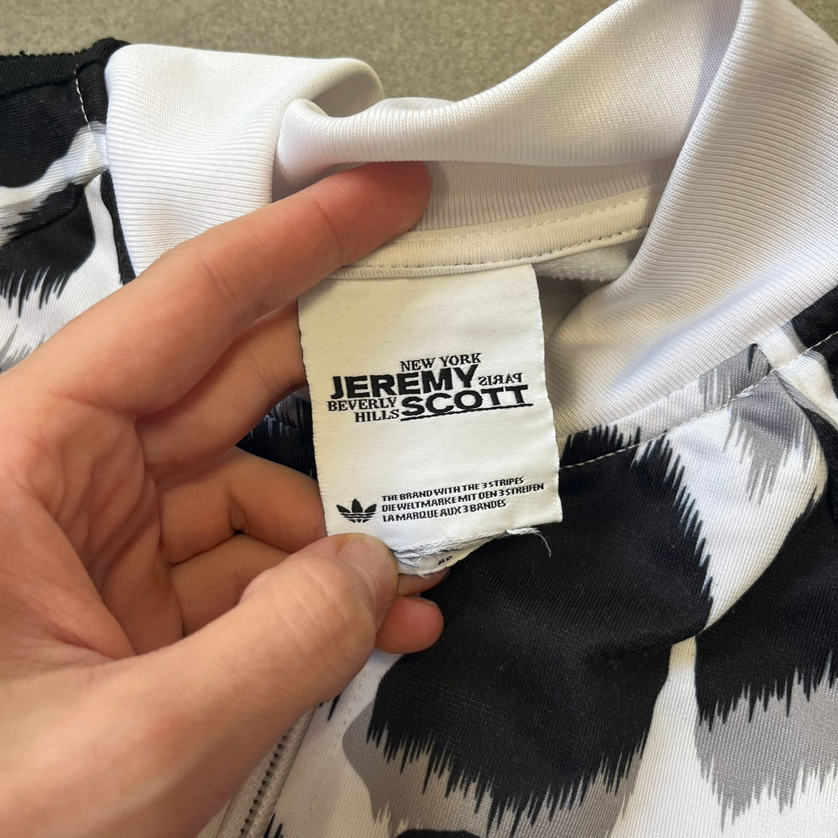 Adidas Jeremy Scott 2015 White Leopard Track Jacket is a white track jacket with a black and gray leopard print, with some of the leopard patches filled in with colorful patterns. Embroidered Adidas stripes run along the sleeves and shoulders, and colorful squiggly Adidas text appears on the chest. The back repeats the same text under a large colorful Trefoil logo, with all colored parts made of mixed patterns and colors.